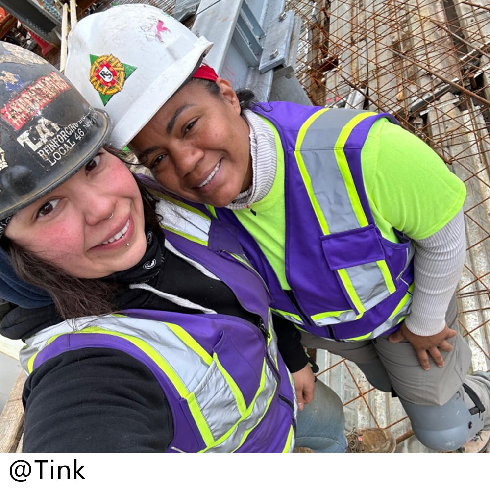 Two individuals in construction attire with hard hats and purple safety vests on a construction site.